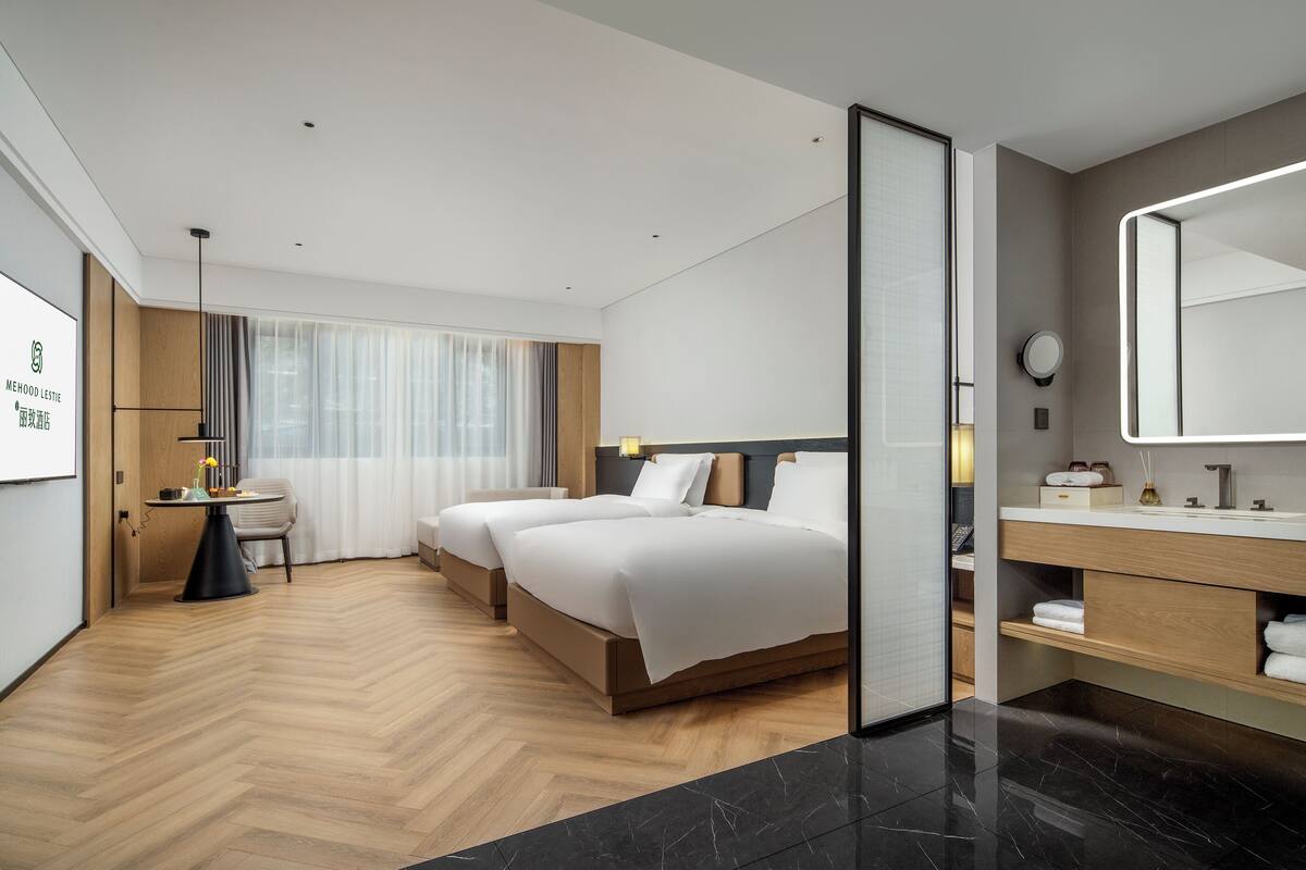 Ein modernes Hotelzimmer mit zwei Betten, einer Sitzecke, einem großen Spiegel und einem Badezimmer mit Waschtisch.