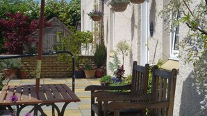 Un banc et une table en bois se trouvent dans un espace jardin, et des jardinières suspendues ornent un bâtiment.