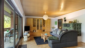 Ein modernes Wohnzimmer mit einer Couch, einem Fernseher, Holzschränken und Blick durch große Fenster.