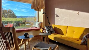 Ein Zimmer mit einem gelben Sofa, einem Holzstuhl, einem kleinen Tisch und einem Fenster mit Blick auf Grünflächen.