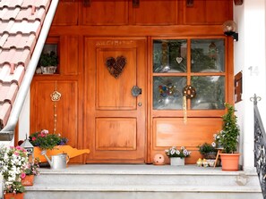 Une porte en bois ornée d’un motif en forme de cœur, une fenêtre décorée de motifs en forme de cœur et des plantes en pot sur le porche.