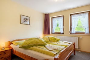 A bedroom with a wooden bed, a nightstand with a lamp, a framed picture on the wall, and windows with curtains.