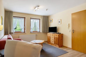A room with a sofa, a small table, a TV, a wooden cabinet, and two windows with curtains.