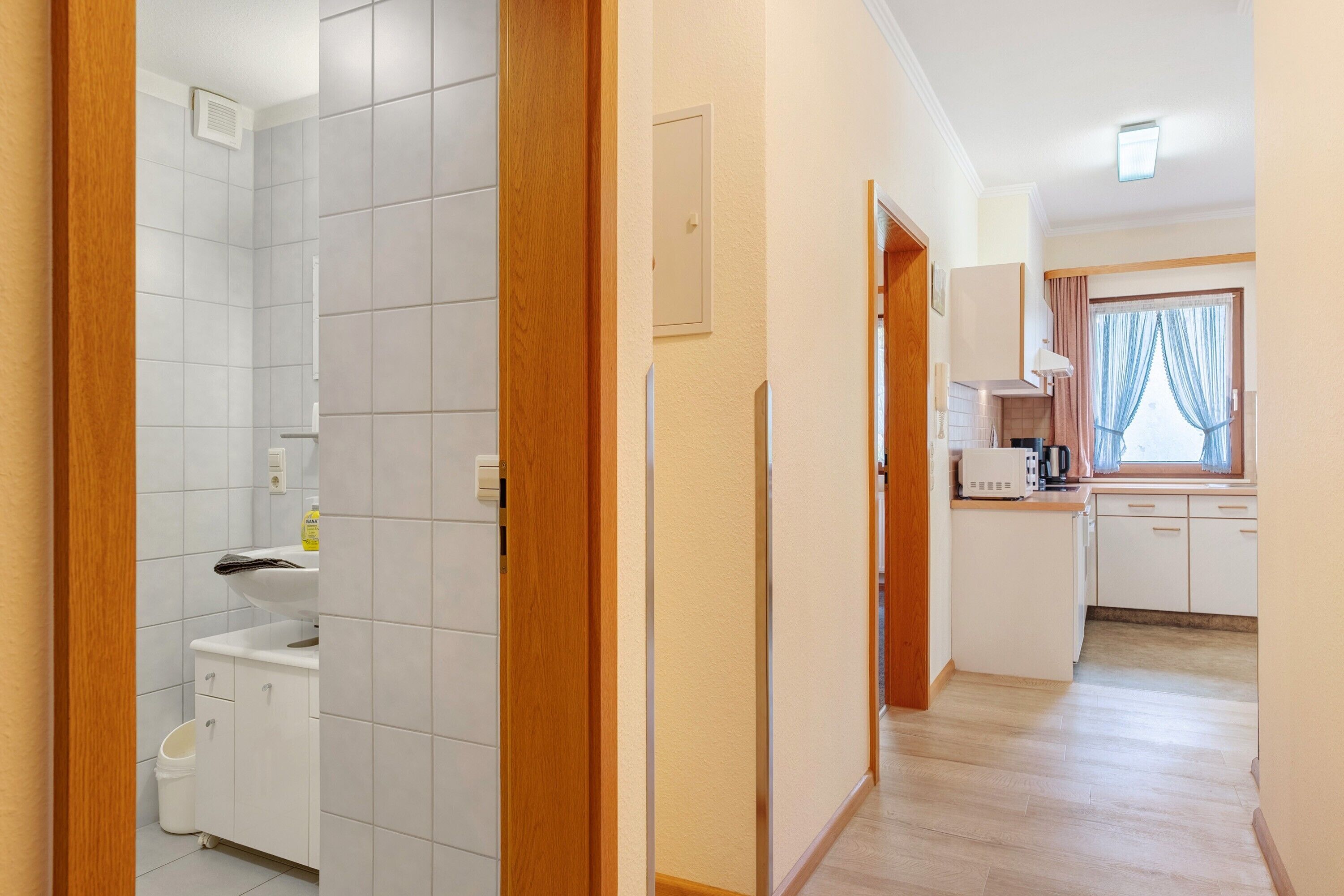A hallway leading to a kitchen with white cabinets, a microwave, and a window with curtains.