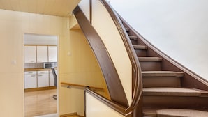 Una escalera de madera con barandilla, que conduce a una zona de cocina con armarios blancos y un microondas.