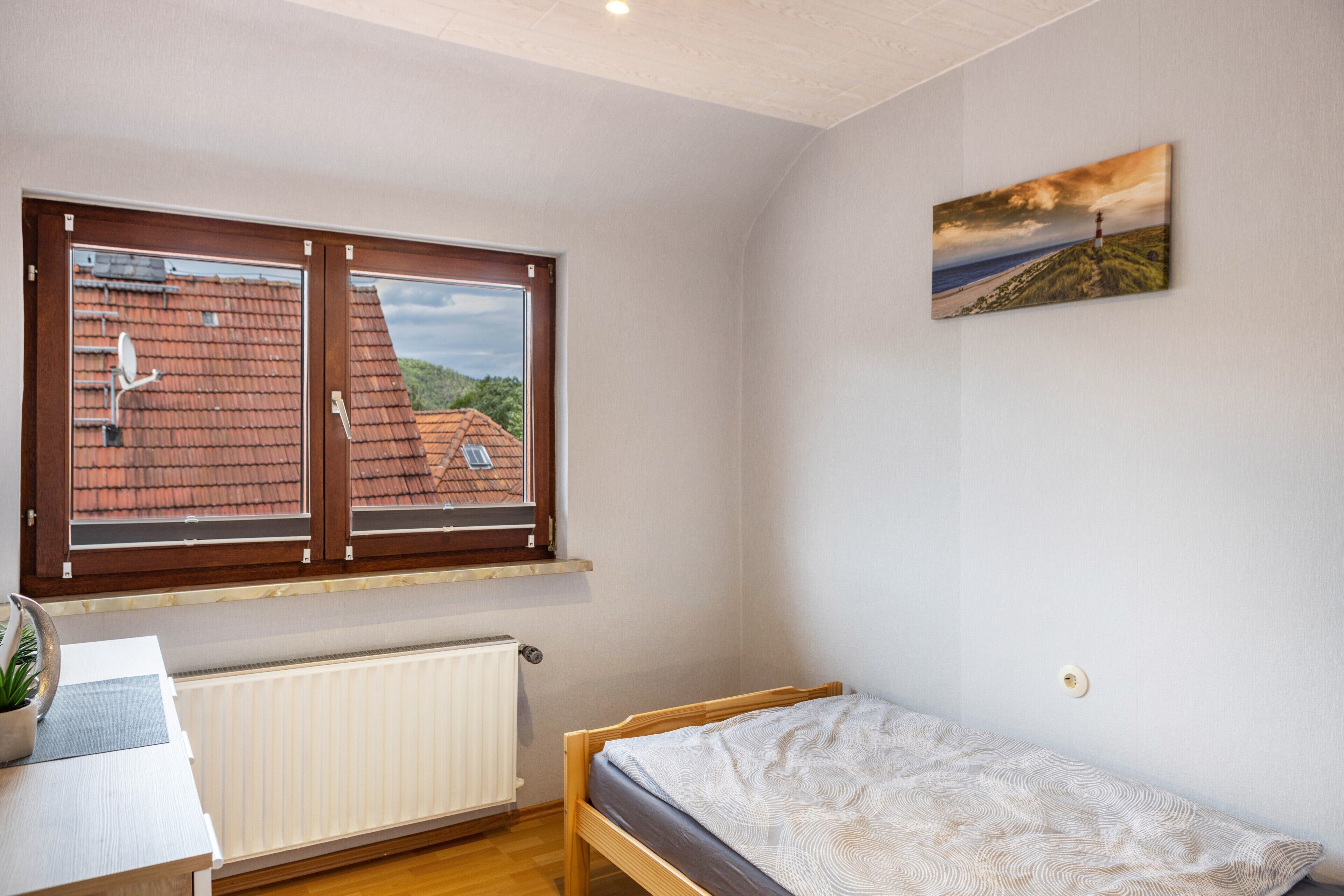 Una camera da letto con un letto di legno, una finestra che si affaccia su un edificio con tetto rosso, un termosifone e un tavolino con una pianta.