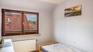 Una camera da letto con un letto di legno, una finestra che si affaccia su un edificio con tetto rosso, un termosifone e un tavolino con una pianta.