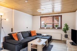A modern living room with a sectional sofa, a wooden coffee table, a TV stand, a potted plant, and a window with a view of buildings.