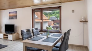 Una habitación moderna con una mesa de comedor, sillas, televisor y un amplio ventanal con vistas a casas.