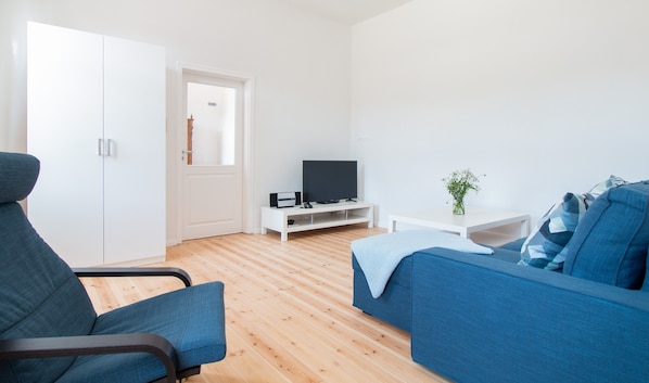 Una sala de estar moderna con un sofá, un sillón, un televisor sobre un mueble blanco y una puerta que da a otra habitación.