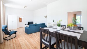 A modern living room with a blue sofa, a dining table with chairs, and a TV in the background. - Apartment 'Schloss Glücksburg' with Lake View, Private Garden and Wi-Fi (Glücksburg (Ostsee))