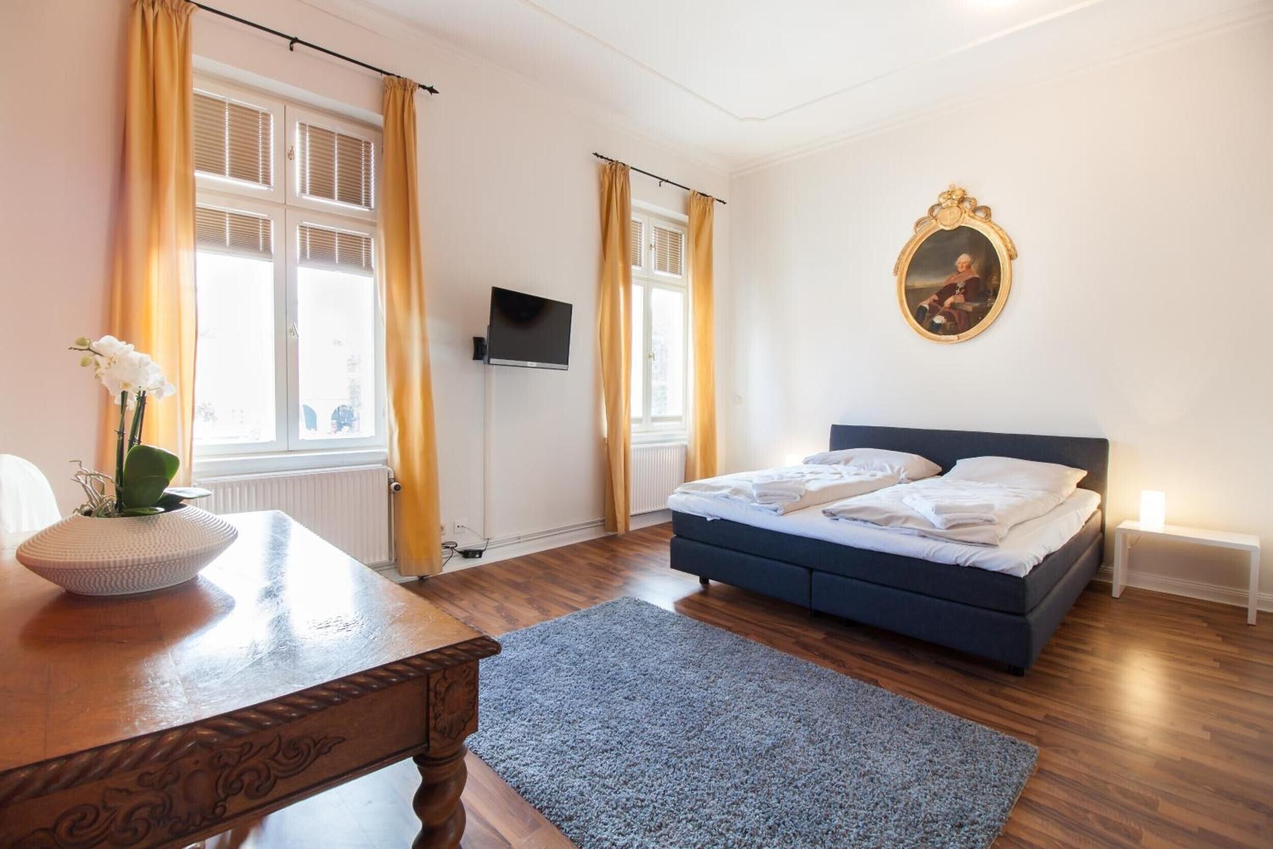 A bedroom with a bed, a TV, a painting, and a table with a vase.