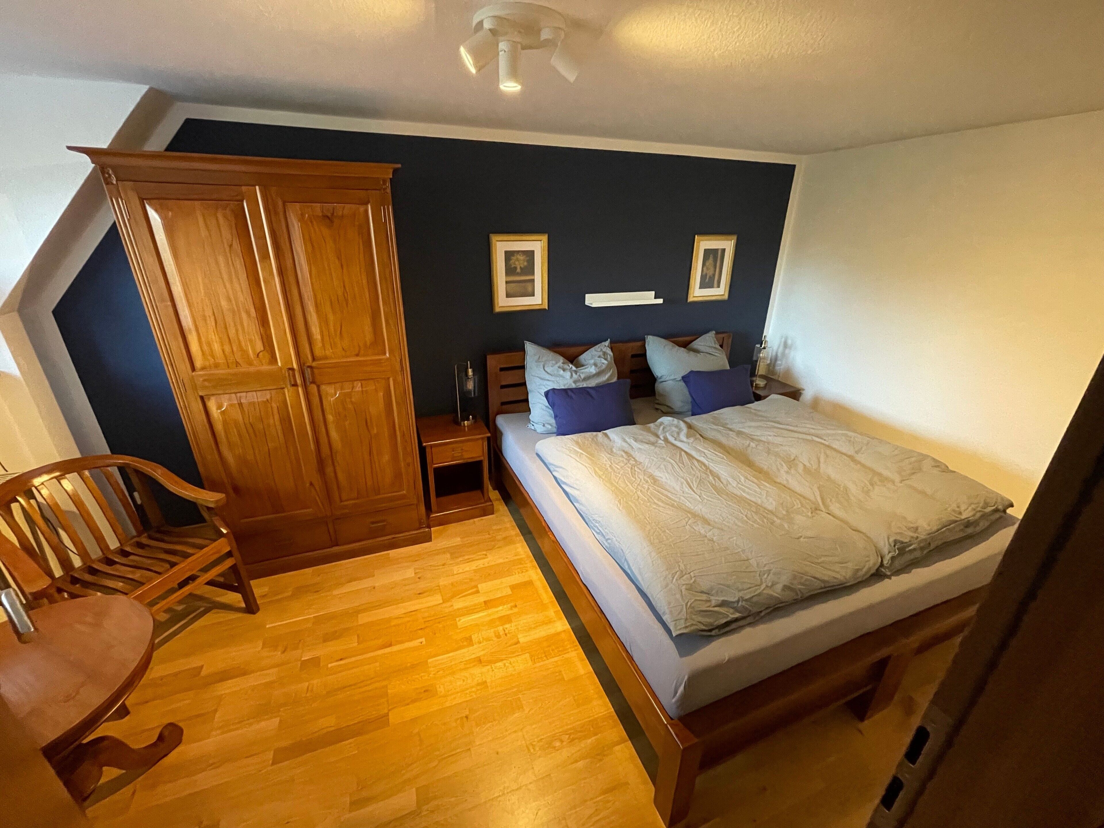 A bedroom with a bed, wooden wardrobe, nightstand, and a chair.