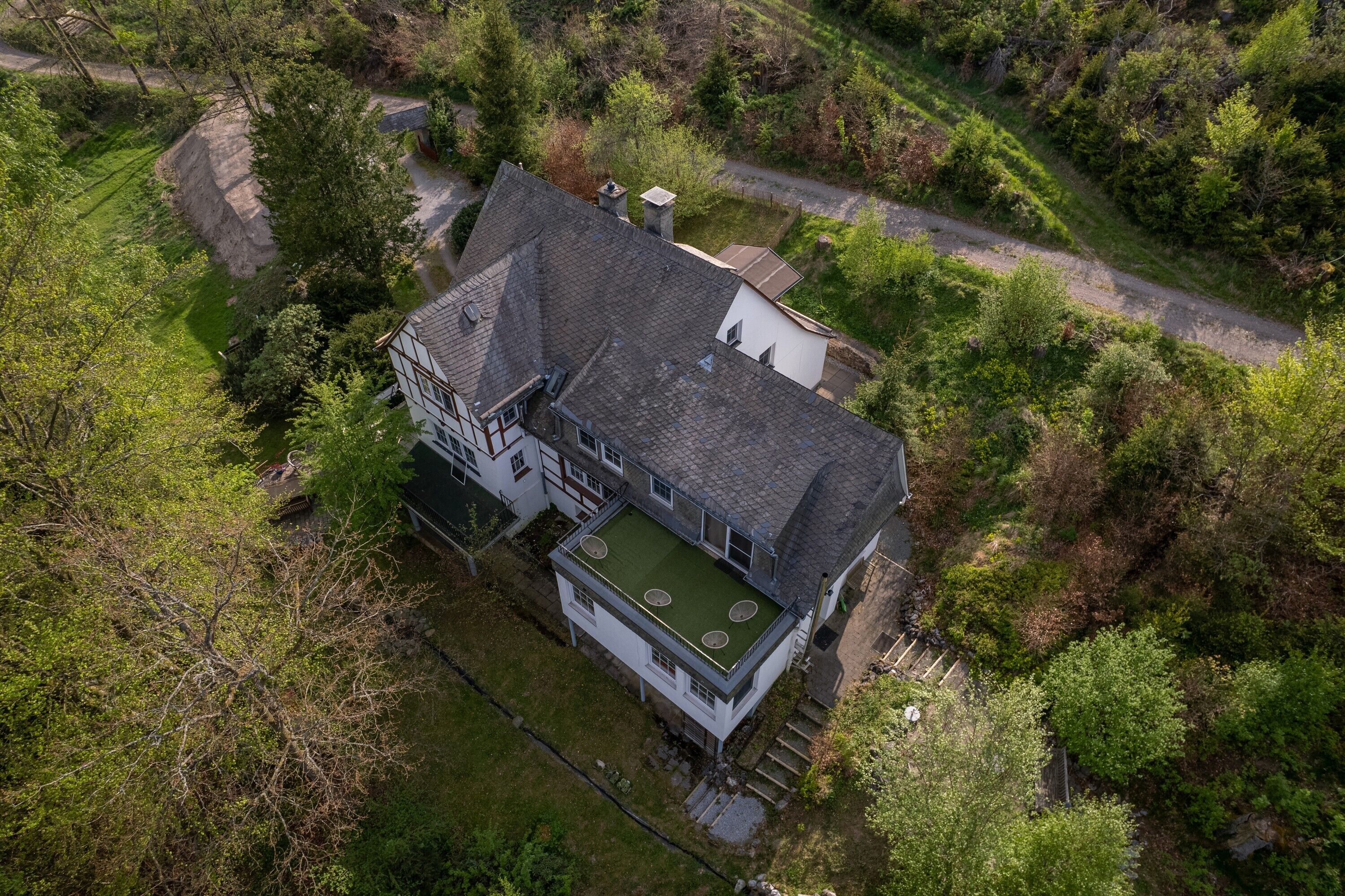 An aerial view of a house with a sloped roof, a green roof extension, and a surrounding wooded area.