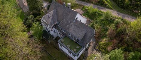An aerial view of a house with a sloped roof, a green roof extension, and a surrounding wooded area.