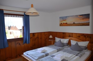 Een slaapkamer met een houten hoofdeinde, een bed met kussens en een deken, een nachtkastje met een lamp en een schilderij van een zonsondergang aan de muur.