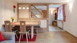 A modern interior with a wooden dining table, chairs, a red rug, a staircase, and a window with checkered curtains.