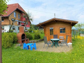 Een houten vakantiehuisje met een bakoven, een terras met een tafel en stoelen, en een bloemrijke tuin.