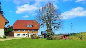 A two-story house with a red roof, a large tree in front, and a small shed in the background.