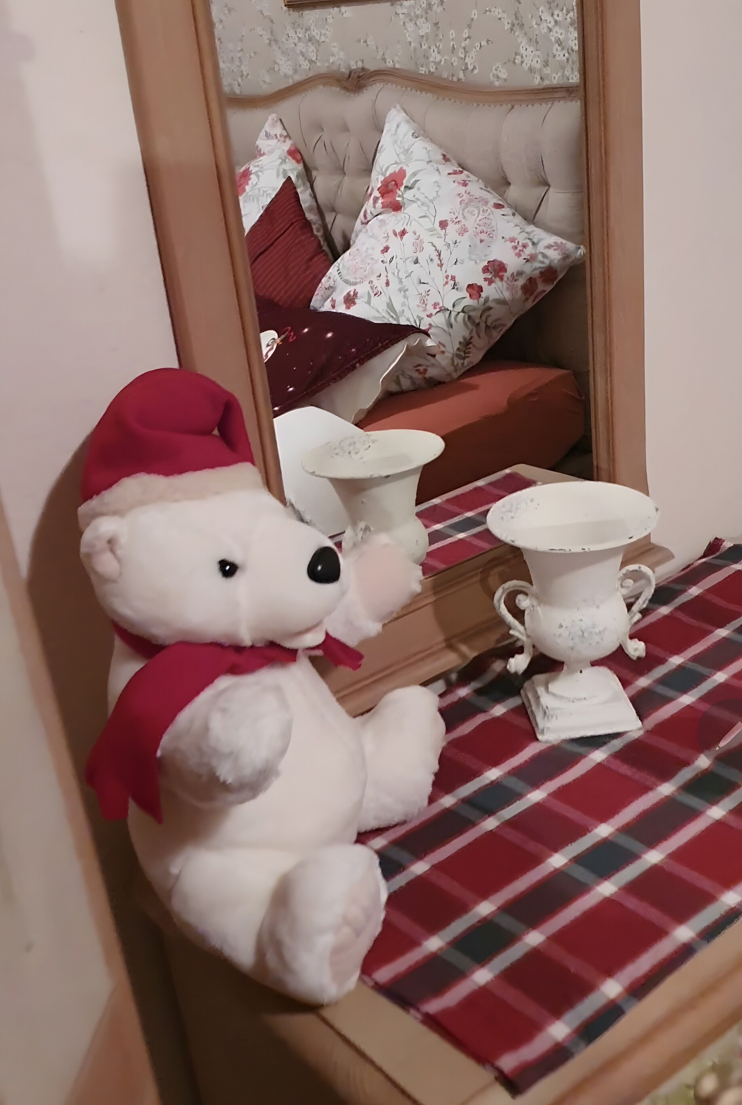 A plush white teddy bear with a red hat sitting on a plaid table next to a white vase.
