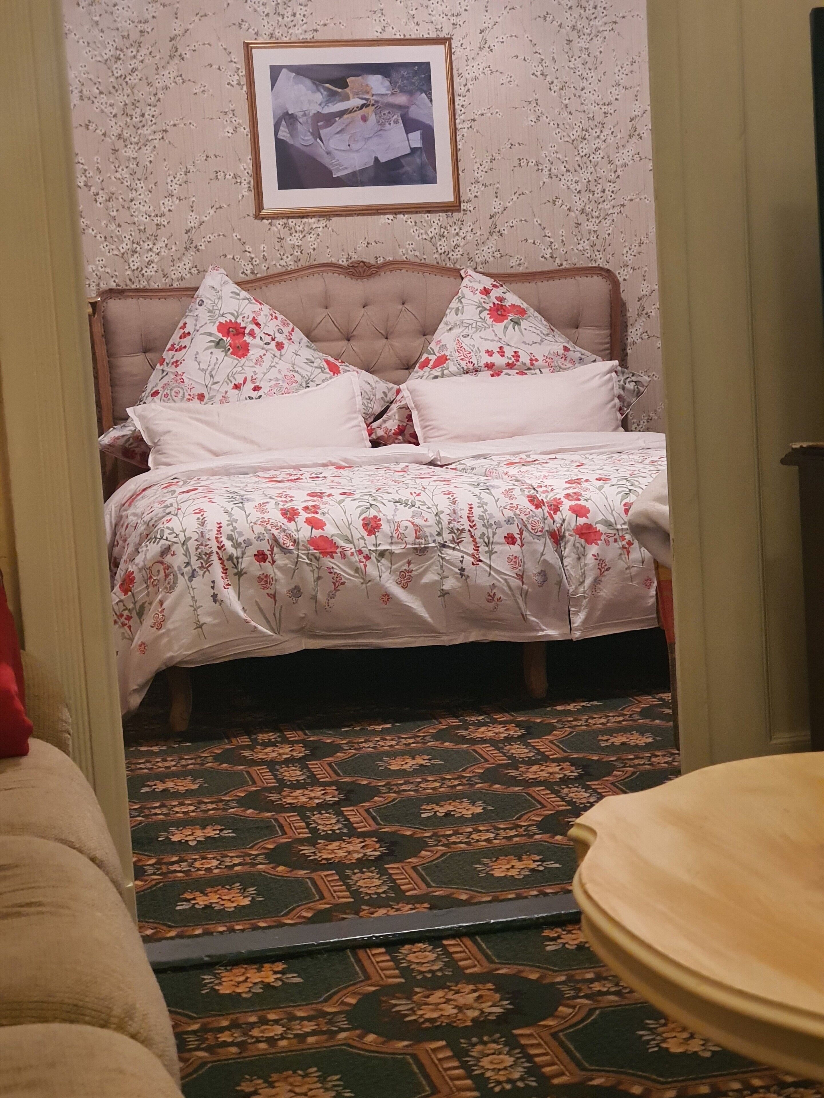 Une chambre à coucher avec une couvre-lit à motifs floraux, une tête de lit capitonnée, une moquette à motifs et un tableau encadré accroché au mur.