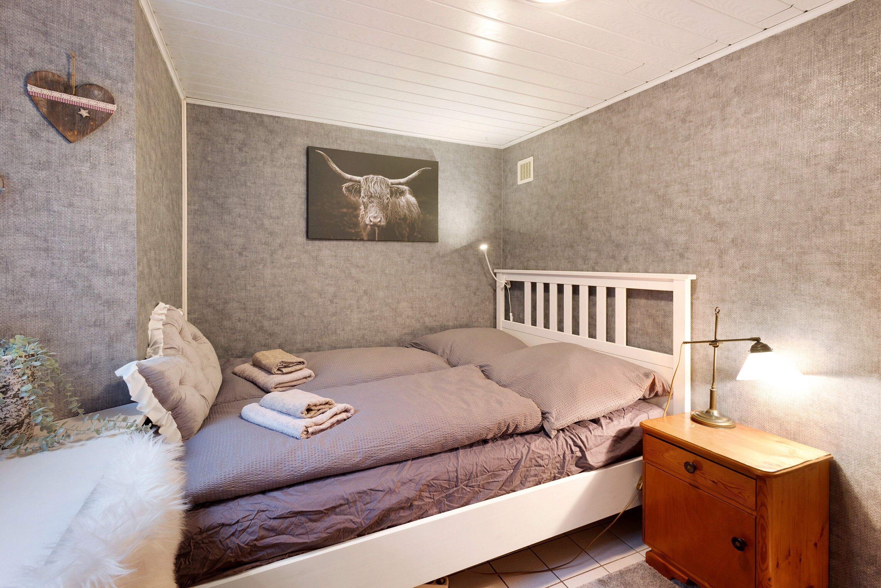 Un dormitorio con cama, almohadas, una mesita de noche con lámpara, un cuadro colgado en la pared y un pequeño mueble auxiliar de madera.