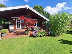 A house with a covered patio, a red maple tree, and a variety of flowers. - Holiday Home 'Rita in Falkenburg' – Relaxation, Nature, WiFi (Hohenwestedt)