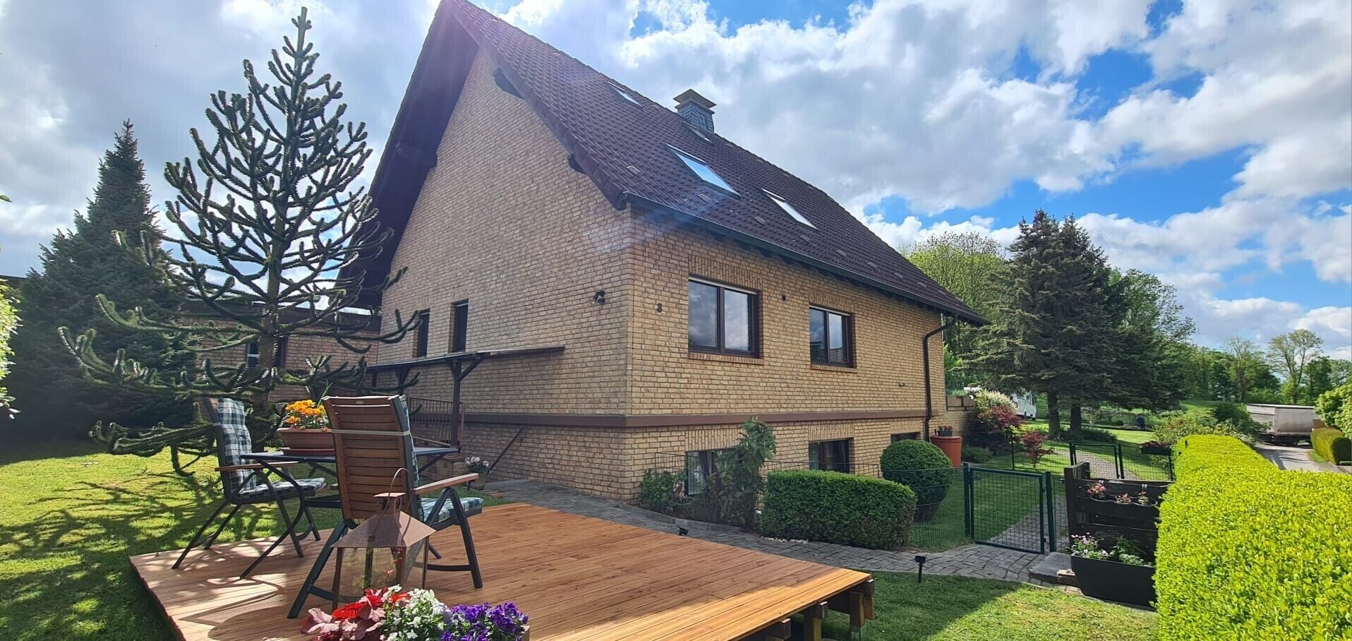 A two-story house with a sloped roof, a brick exterior, and a fenced backyard with a wooden deck and outdoor seating.