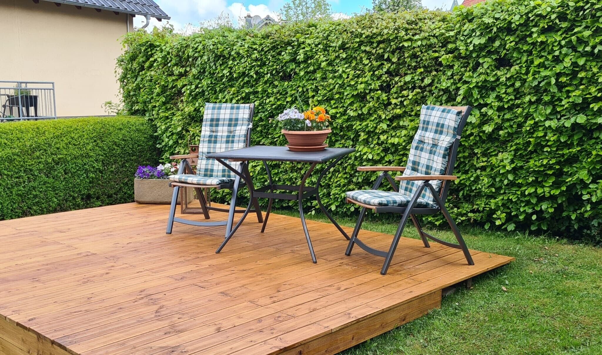 Una terraza de madera con dos sillas y una mesa, rodeada por un seto.