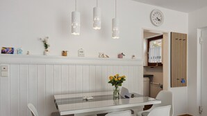A dining area with a glass table, white chairs, a vase with yellow flowers, pendant lights, and a clock on the wall.