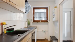 A compact kitchen with a sink, stove, and cabinets, a window with curtains, and a small shelf with bottles.