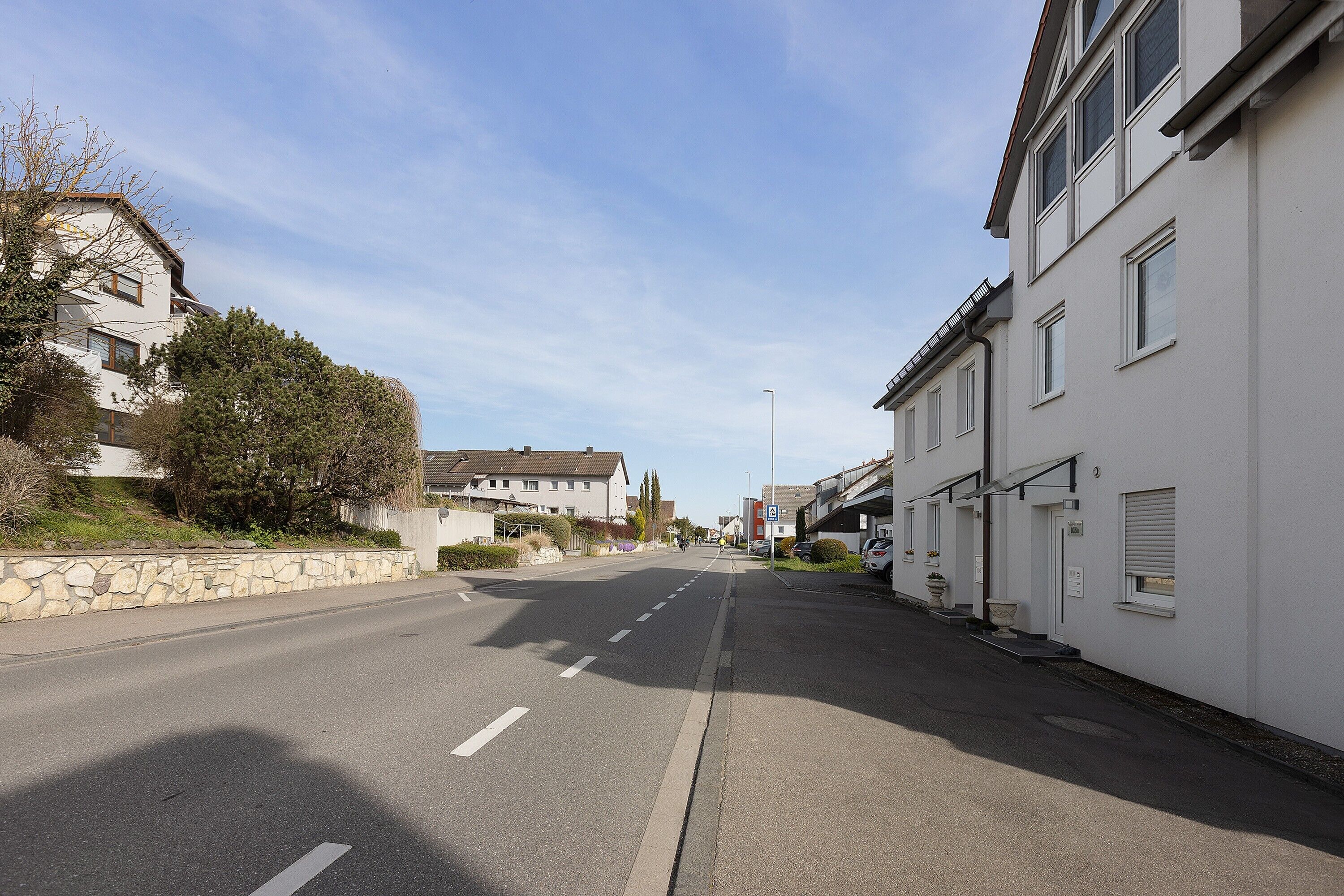 Une rue résidentielle tranquille, bordée de maisons, avec un ciel dégagé et un mur de pierre sur le côté gauche.