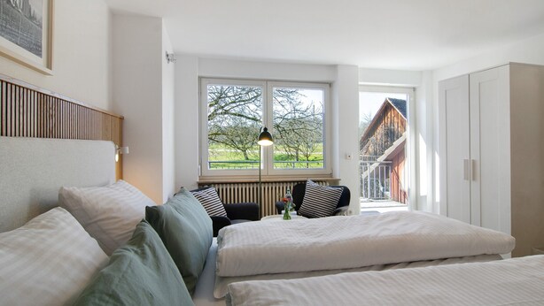 Une chambre avec deux lits, une fenêtre donnant sur un immeuble et des arbres, un radiateur et une petite table avec un vase.