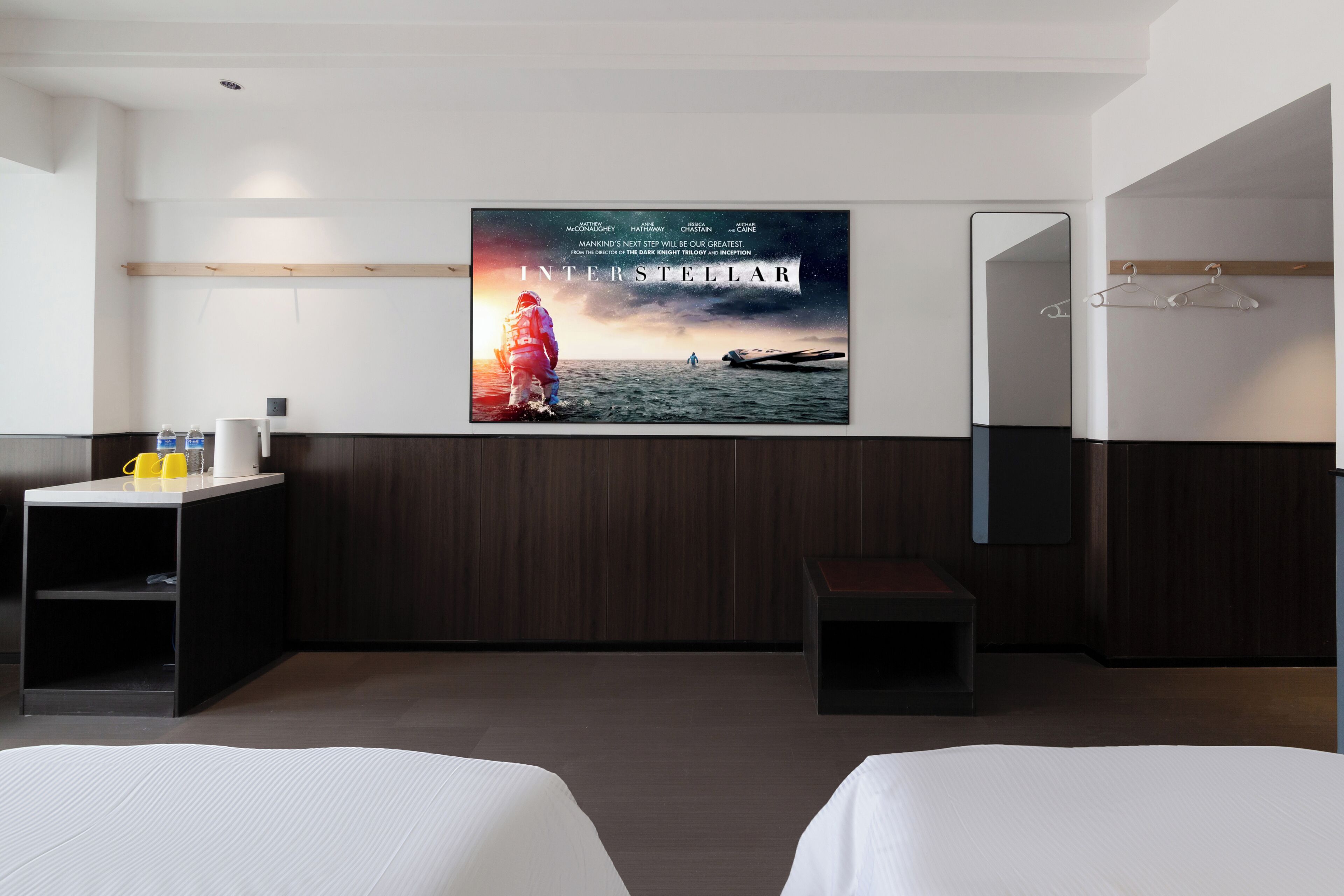 Ein Hotelzimmer mit einem großen Poster von 'Interstellar' an der Wand, einem Bett, einem kleinen Tisch mit einer Tasse und einer Flasche, einem Spiegel und einem Holzregal.