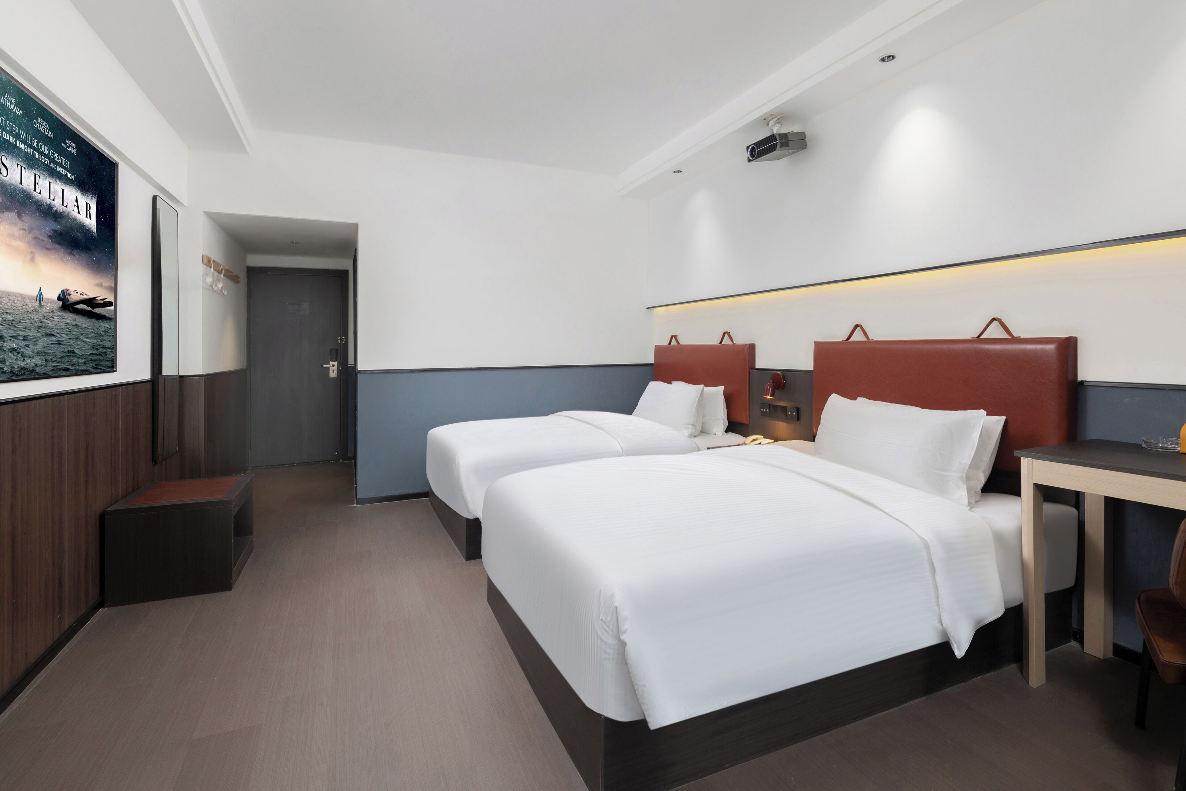 A hotel room with two beds, a desk, and a seating area. There is a poster on the wall and a door leading to another room.