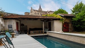 Un espace aménagé au bord de la piscine, avec une terrasse carrelée, une piscine et un bâtiment dont l’arrière-plan est dominé par une église.
