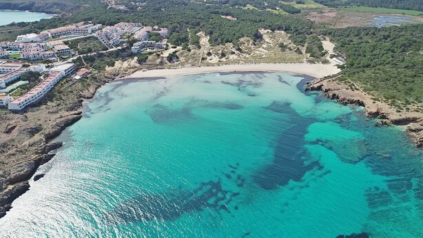 Vista aérea de una zona costera con una playa de arena y aguas turquesas cristalinas. Se ven edificios de varios pisos al fondo, lo que sugiere una zona residencial o turística.