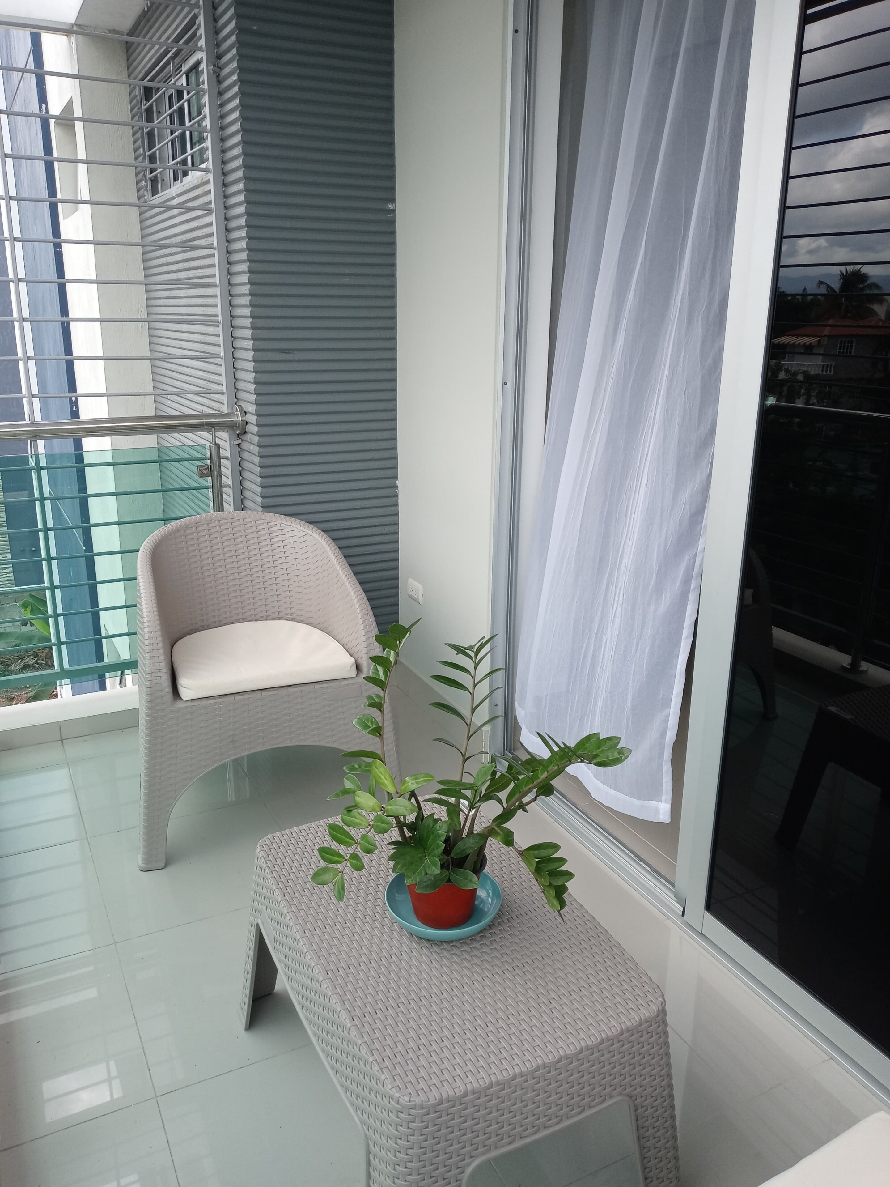 A balcony with a wicker chair, a small table with a potted plant, and a window with a white curtain.