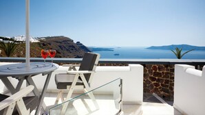 A terrace with a table, chairs, and a glass table. There are two glasses on the table. The terrace overlooks a body of water and a mountainous landscape. - Two-Bedroom Caldera View (Thira - Santorini)