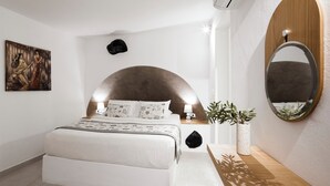 A modern bedroom with a large bed, a wall-mounted mirror, a painting, and a potted plant. - Two-Bedroom Caldera View (Thira - Santorini)