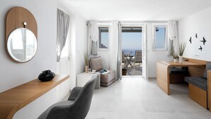 A modern, well-lit room with a wooden desk, a grey chair, a small sofa, and a view of the sea through open doors. - Two-Bedroom Caldera View (Thira - Santorini)