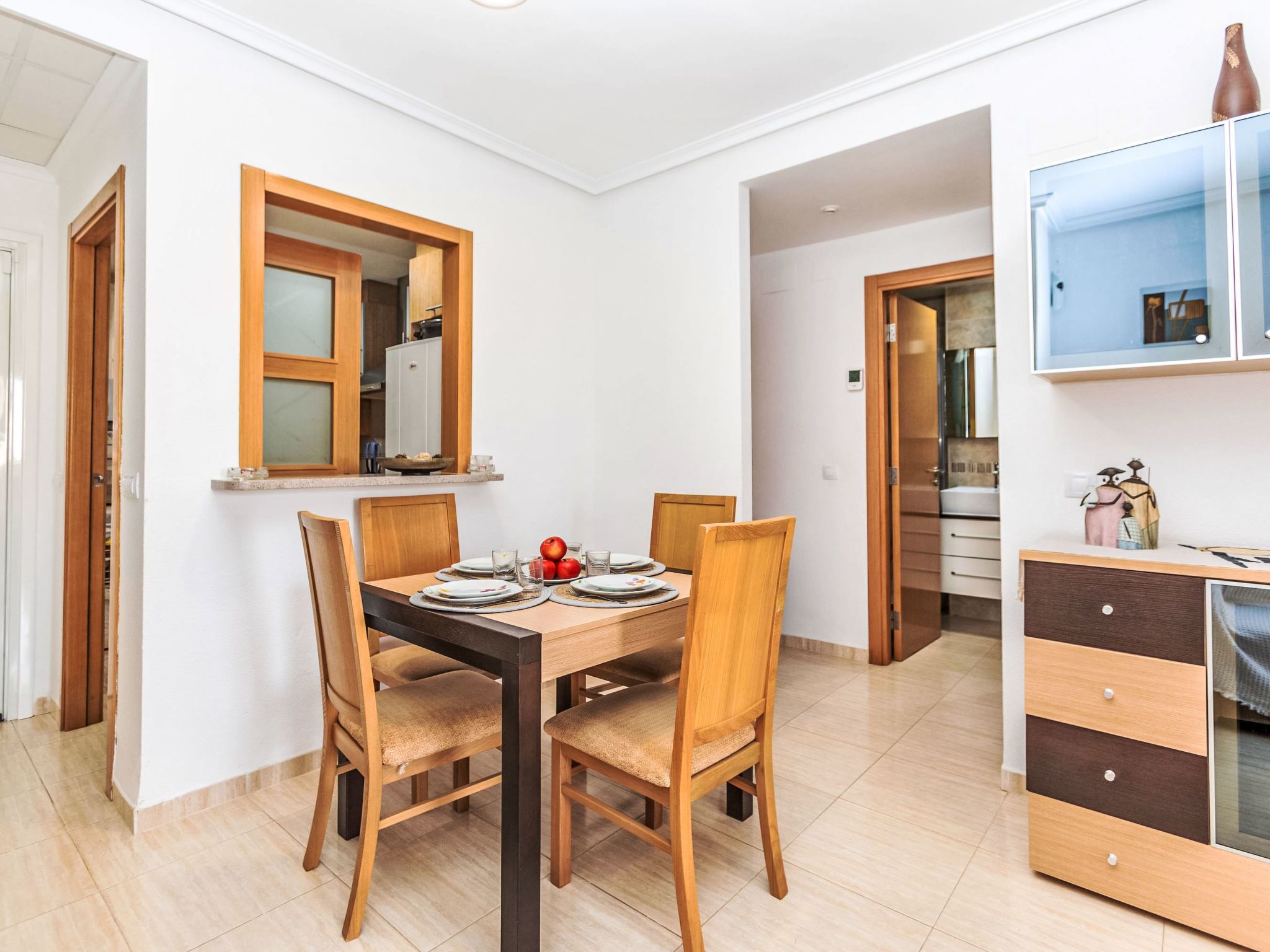 A dining area with a wooden table and chairs, a window with a wooden frame, and a kitchen area with cabinets and a refrigerator.