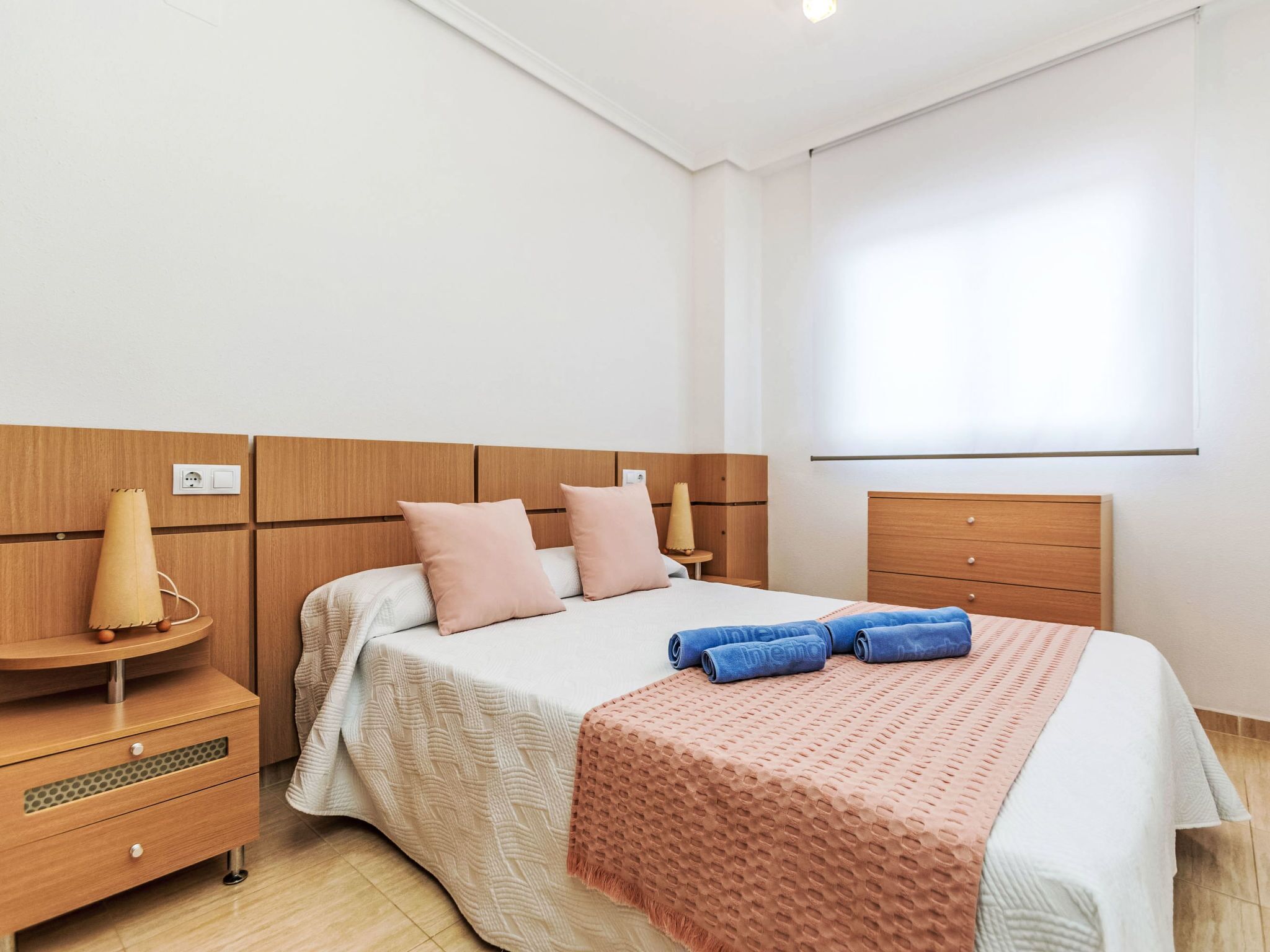 Une chambre à coucher comprenant un lit, une table de chevet, une commode en bois et une fenêtre avec des stores.