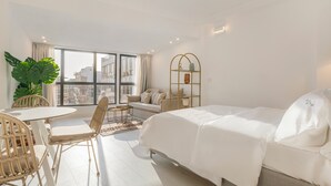 Habitación de hotel moderna con cama, sofá, mesa de comedor con sillas, ventana grande y planta en maceta.