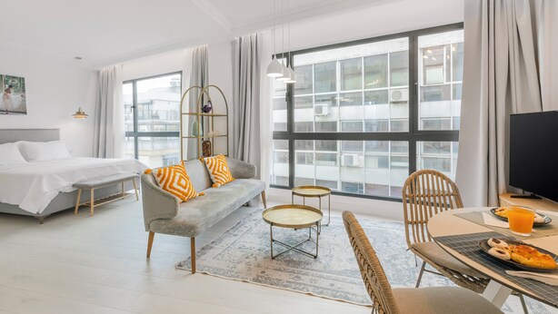 Una habitación de hotel moderna con una cama, un sofá gris, una mesa de comedor con sillas y un ventanal con cortinas.