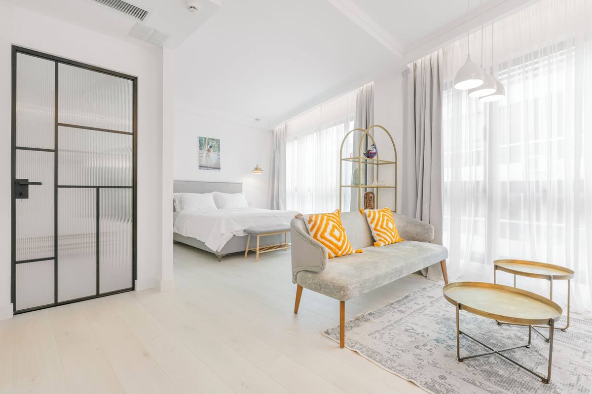 Moderni hotellihuone, jossa on sänky, sohva, kaksi sivupöytää ja suuri ikkuna, jossa on verhot.