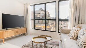 Una sala moderna con un televisor de pantalla plana grande sobre un mueble de madera, un sofá con cojines estampados y un ventanal con vista a la ciudad.