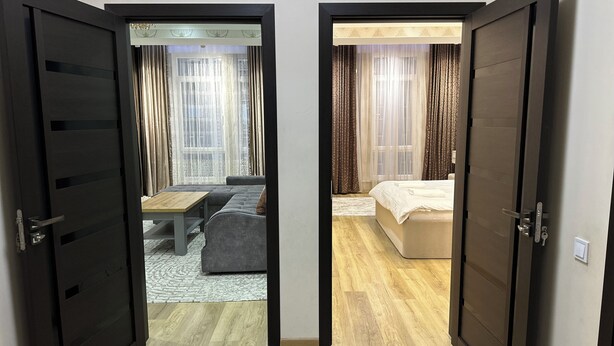 Hotellihuone, jossa on sänky, sohva, pieni pöytä ja ikkunasta verhojen läpi avautuva näkymä ulos.