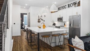 Een moderne keuken met witte kasten, een centraal eiland met barkrukken, een roestvrijstalen koelkast, en een muur met 'MUSIC ROW' er bovenop.