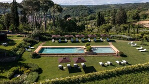 Zona de piscina con sillones de descanso y sombrillas, rodeada de césped cuidado y árboles.